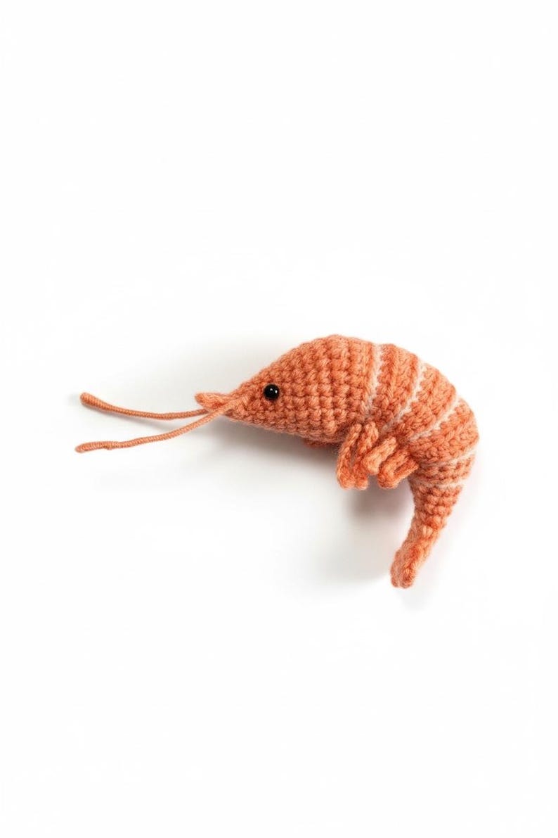 May include: A handmade, orange crochet shrimp with white stripes and black eyes. The shrimp is lying on a white surface. The shrimp's antennae and legs are visible. This item is a unique, handcrafted piece.