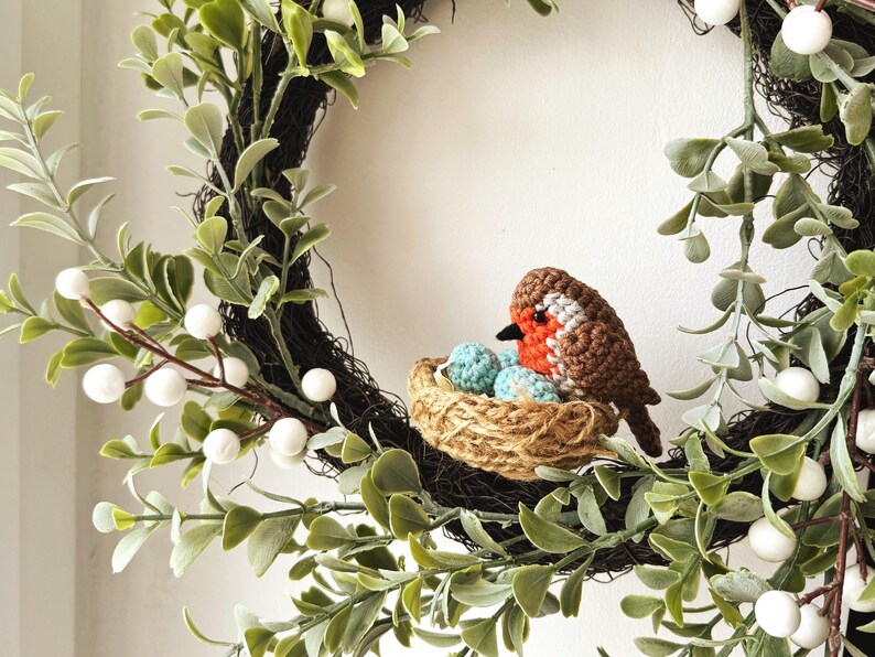 May include: A handcrafted wreath featuring a crocheted robin sitting in a nest with blue eggs. The wreath is adorned with green leaves, white berries, and a dark brown base, creating a charming spring or Easter decoration.
