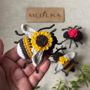 May include: A crocheted bee ornament featuring a yellow and black striped body, white tail, and light grey wings, adorned with a yellow sunflower on its back. Nearby are a black spider with a red flower and another smaller crocheted bee.