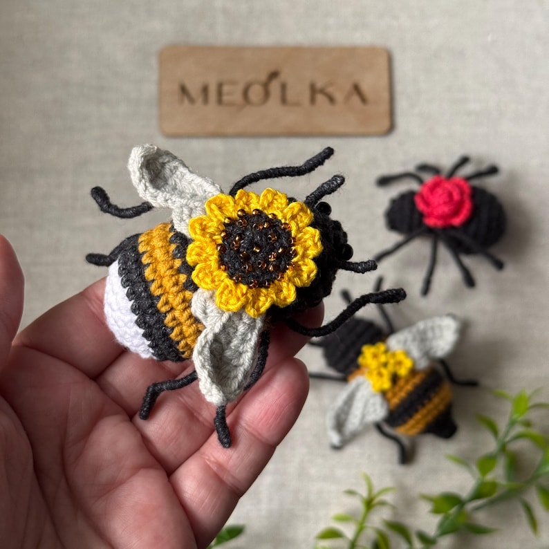 May include: A crocheted bee ornament featuring a yellow and black striped body, white tail, and light grey wings, adorned with a yellow sunflower on its back. Nearby are a black spider with a red flower and another smaller crocheted bee.
