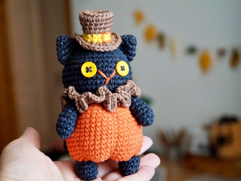 May include: A handmade crochet cat doll with a pumpkin-shaped body and a top hat. The cat is dark blue with yellow button eyes and a brown ruffled collar. The pumpkin body is orange. A charming Halloween decoration.