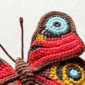 May include: A crocheted butterfly applique featuring intricate yarn work in shades of brown, red, yellow, black, and light blue. The design includes two long, thin antennae and detailed circular patterns on the wings created with textured stitches.