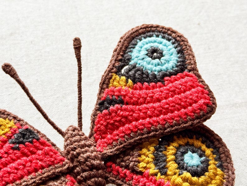 May include: A crocheted butterfly applique featuring intricate yarn work in shades of brown, red, yellow, black, and light blue. The design includes two long, thin antennae and detailed circular patterns on the wings created with textured stitches.