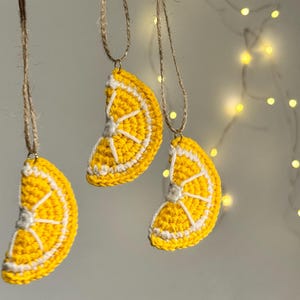 May include: Three handmade crochet lemon slice ornaments hanging from twine. The ornaments are yellow with white details, resembling a lemon slice. The background is a soft, neutral color with a string of fairy lights.