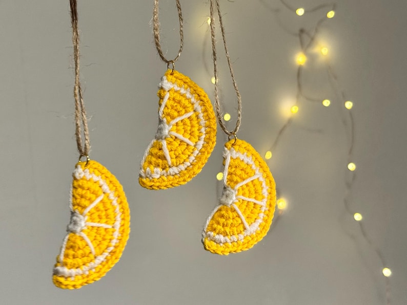May include: Three handmade crochet lemon slice ornaments hanging from twine. The ornaments are yellow with white details, resembling a lemon slice. The background is a soft, neutral color with a string of fairy lights.