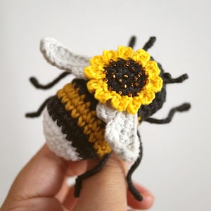 May include: A crocheted bee figurine featuring a black and yellow striped body with a white tail section. The bee has light grey wings and a bright yellow sunflower with a dark brown centre attached to its back, along with thin black legs.