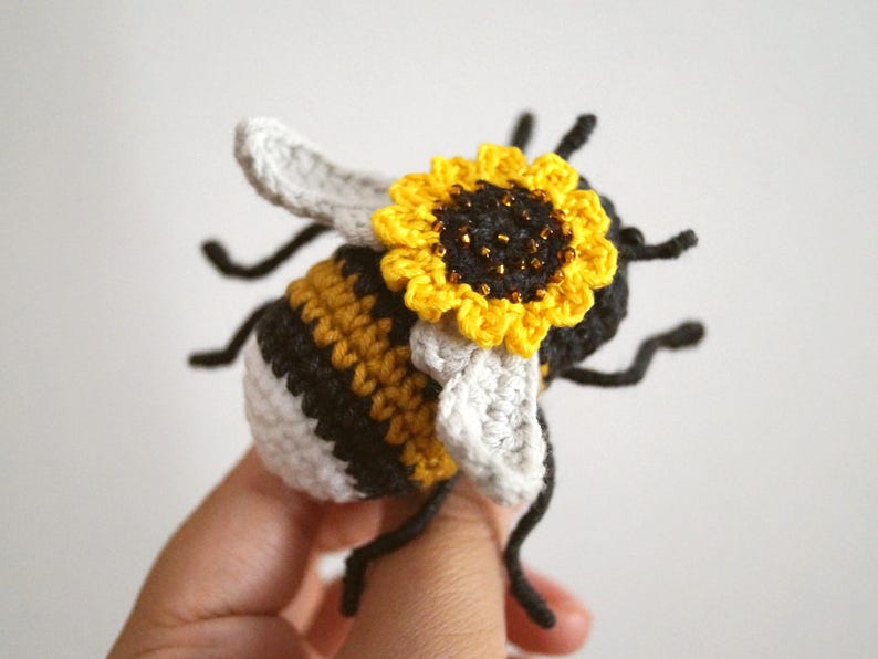 May include: A crocheted bee figurine featuring a black and yellow striped body with a white tail section. The bee has light grey wings and a bright yellow sunflower with a dark brown centre attached to its back, along with thin black legs.
