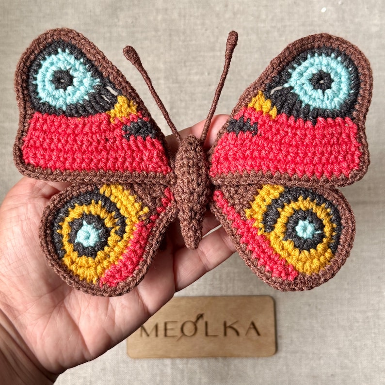 May include: A crocheted butterfly applique featuring a brown body and antennae. The wings display a vibrant pattern of red, yellow, dark gray, and light blue yarn. The butterfly is crafted with detailed stitching and has a symmetrical design.