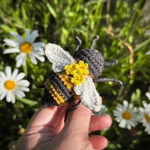 May include: A crocheted bee toy featuring a striped yellow and dark gray body, light gray wings, and a cluster of small yellow flowers attached to the back. The bee has dark gray antennae and legs, with a textured yarn construction throughout.