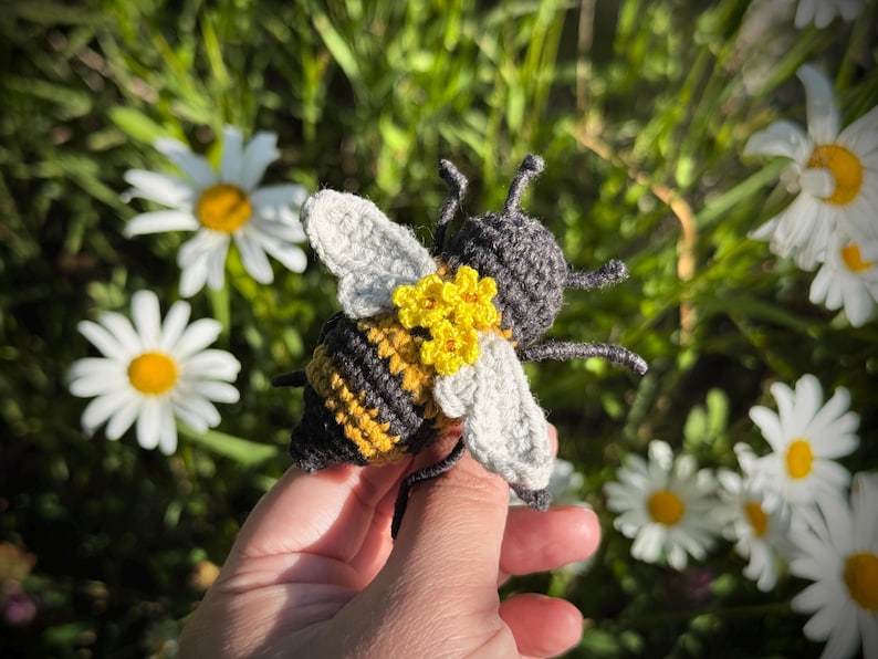 May include: A crocheted bee toy featuring a striped yellow and dark gray body, light gray wings, and a cluster of small yellow flowers attached to the back. The bee has dark gray antennae and legs, with a textured yarn construction throughout.
