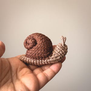 Puede incluir: Un pequeño caracol de ganchillo hecho a mano con una concha en espiral de color marrón oscuro y un cuerpo de color beige claro. El caracol tiene dos pequeñas antenas erguidas en la cabeza y está tejido con puntos visibles y texturizados.