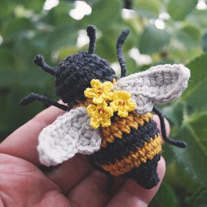 May include: A crocheted bee toy featuring a black and yellow striped body, light gray wings, and three small yellow flowers with brown centers attached to the back. The bee has black antennae and thin black legs extending from the sides.