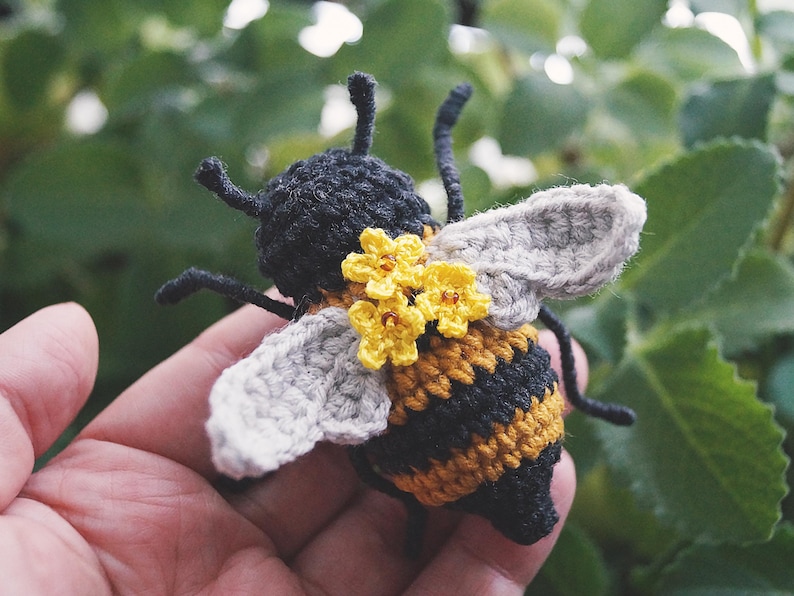 May include: A crocheted bee toy featuring a black and yellow striped body, light gray wings, and three small yellow flowers with brown centers attached to the back. The bee has black antennae and thin black legs extending from the sides.