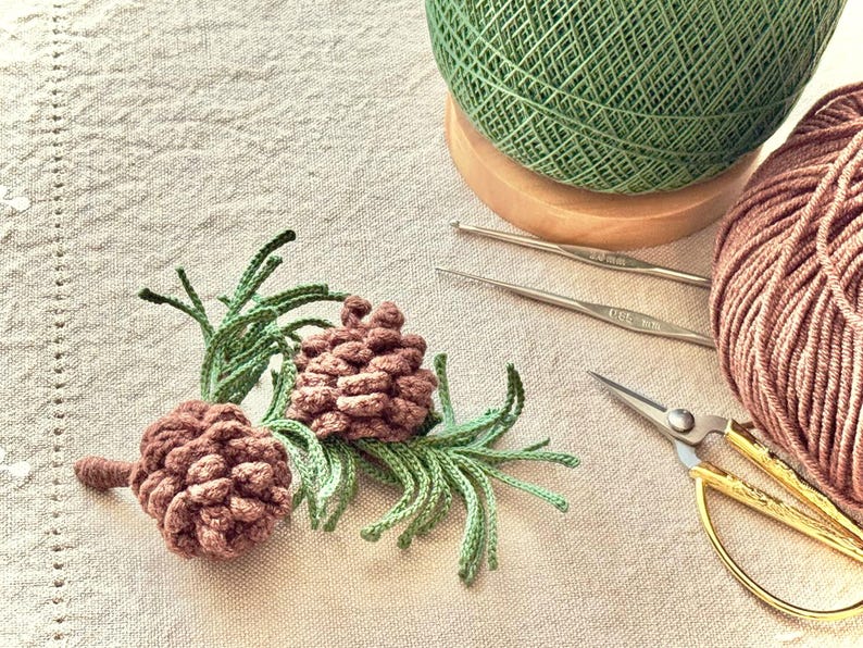 Op de afbeelding: Een close-up van twee gehaakte dennenappels met groene naaldachtige details. Een bol groen garen staat op een houten voet, met een bol bruin garen, twee haaknaalden en een gouden schaar.