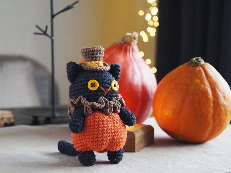May include: A handmade crochet cat doll wearing a brown top hat, ruffled collar, and orange pants. The cat has yellow button eyes and a black body. Two pumpkins are in the background, one orange and one red.