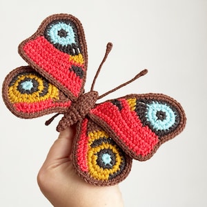 May include: A handcrafted crochet butterfly featuring a brown body and antennae, with wings displaying vibrant patterns of red, mustard yellow, light blue, and black. The intricate yarn work creates a detailed, textured appearance on the wings.