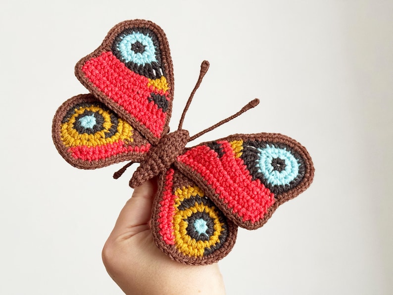 May include: A handcrafted crochet butterfly featuring a brown body and antennae, with wings displaying vibrant patterns of red, mustard yellow, light blue, and black. The intricate yarn work creates a detailed, textured appearance on the wings.