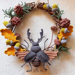 Op de afbeelding: Een decoratieve krans van bruine takken versierd met gehaakte gele eikenbladeren, bruine dennenappels, kleine paddenstoelen en eikels. Een grote, donkergrijze gehaakte vliegend hert is in het midden geplaatst boven een houten label met MEOLKA.