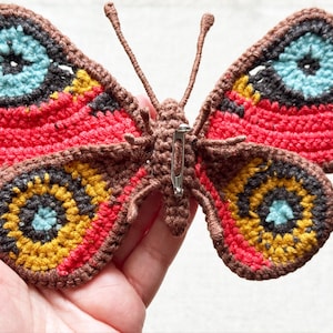 May include: A crocheted butterfly brooch featuring a brown body and antennae with wings in shades of red, yellow, dark brown, and light blue. The back of the butterfly displays a silver-toned metal pin mechanism attached to the central brown body.