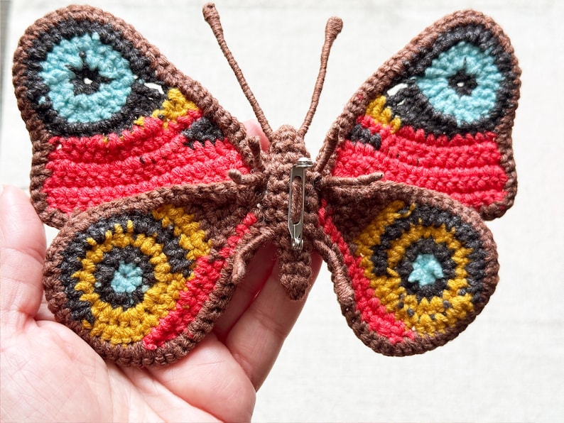 May include: A crocheted butterfly brooch featuring a brown body and antennae with wings in shades of red, yellow, dark brown, and light blue. The back of the butterfly displays a silver-toned metal pin mechanism attached to the central brown body.