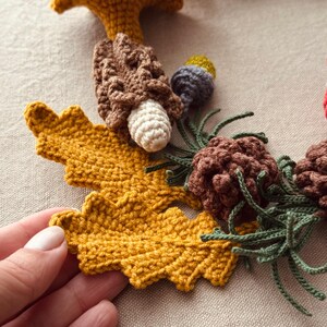 Pode incluir: Grinalda de outono de croch&ecirc; com uma grande folha amarelo mostarda, cogumelos castanhos, pinhas e galhos verdes. A grinalda artesanal tem um tema r&uacute;stico e natural.