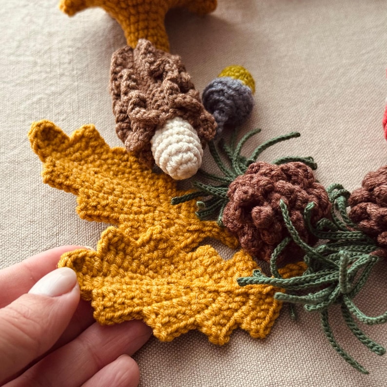 Pode incluir: Grinalda de outono de croch&ecirc; com uma grande folha amarelo mostarda, cogumelos castanhos, pinhas e galhos verdes. A grinalda artesanal tem um tema r&uacute;stico e natural.