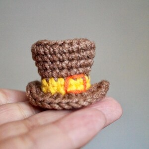 May include: A miniature brown crochet top hat with a yellow band and orange buckle. The hat is handmade and appears to be a decorative item or accessory. The hat is held in a person's hand.