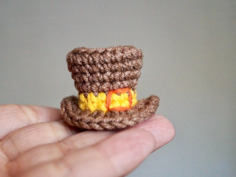 May include: A miniature brown crochet top hat with a yellow band and orange buckle. The hat is handmade and appears to be a decorative item or accessory. The hat is held in a person's hand.
