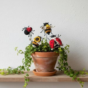 May include: Four crocheted insect ornaments on thin metal stakes inserted into a terracotta pot containing a trailing green plant. The ornaments include a black spider with a red flower, two yellow and black striped bees, and a red ladybug.