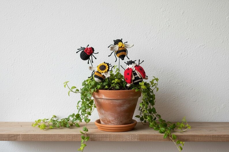 May include: Four crocheted insect ornaments on thin metal stakes inserted into a terracotta pot containing a trailing green plant. The ornaments include a black spider with a red flower, two yellow and black striped bees, and a red ladybug.