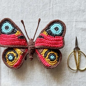 May include: A crocheted butterfly with brown, red, yellow, and light blue yarn, featuring two antennae. A wooden ruler shows the butterfly is approximately 11 cm wide. Beside it are gold-toned embroidery scissors and a thin metal crochet hook.