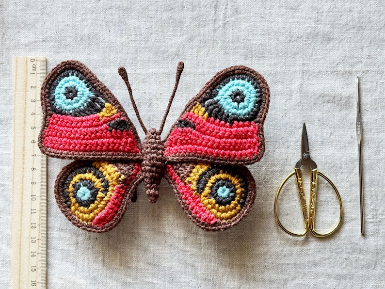 May include: A crocheted butterfly with brown, red, yellow, and light blue yarn, featuring two antennae. A wooden ruler shows the butterfly is approximately 11 cm wide. Beside it are gold-toned embroidery scissors and a thin metal crochet hook.