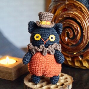 May include: A handmade crochet black cat doll wearing an orange outfit, a brown ruffled collar, and a brown top hat with yellow trim. The cat has yellow button eyes and is standing on a wooden slice.