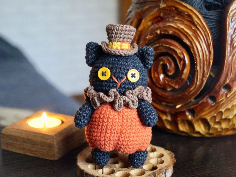 May include: A handmade crochet black cat doll wearing an orange outfit, a brown ruffled collar, and a brown top hat with yellow trim. The cat has yellow button eyes and is standing on a wooden slice.