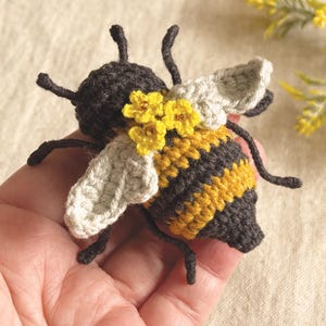 May include: A crocheted bee figurine featuring a dark brown head and abdomen with yellow stripes. The bee has two light beige crocheted wings and is decorated with three small yellow crocheted flowers on its back, complete with thin black antennae and legs.