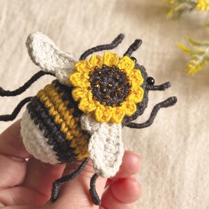 May include: A crocheted bee figurine featuring a striped yellow, black, and white body with off-white wings. A small, bright yellow sunflower with a dark centre and subtle gold bead accents is attached to the top of the bee's head.
