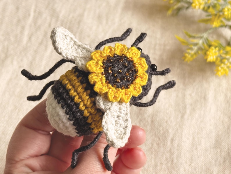 May include: A crocheted bee figurine featuring a striped yellow, black, and white body with off-white wings. A small, bright yellow sunflower with a dark centre and subtle gold bead accents is attached to the top of the bee's head.