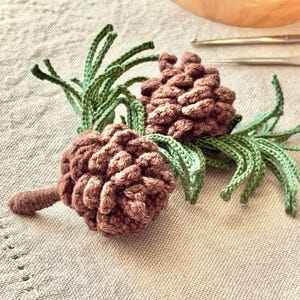 Op de afbeelding: Twee gehaakte dennenappels in een warme bruine kleur, met groene naald details. De handgemaakte items worden getoond op een neutrale stof. Het ambachtelijke werk doet denken aan natuurlijke vormen.