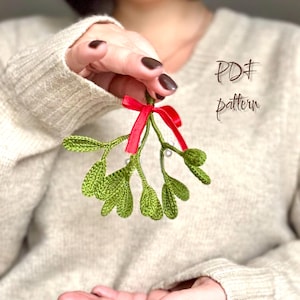 Può includere: Un vischio verde all'uncinetto con un fiocco di nastro rosso e piccoli accenti di perle bianche. Il testo "PDF pattern" è scritto in corsivo. Questo articolo fatto a mano è perfetto per le decorazioni natalizie.