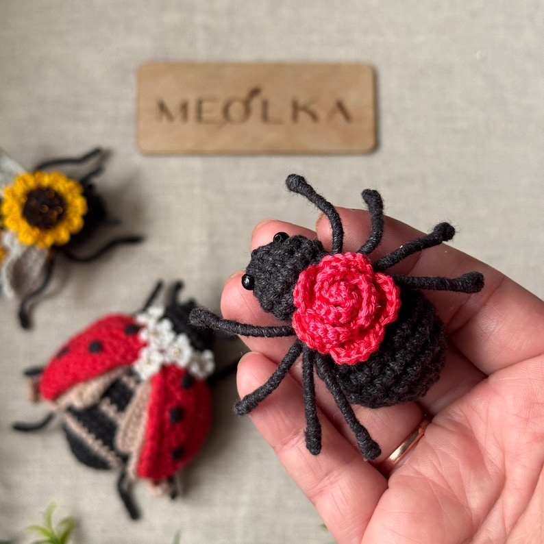 May include: A black crocheted spider with a bright red crocheted rose on its back, featuring small black bead eyes. Nearby are a red and black crocheted ladybug and a yellow crocheted sunflower. A wooden tag with the text MEOLKA is visible.