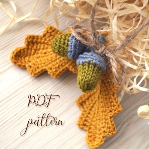 Op de afbeelding: Handgebreide herfstdecoratie met een mosterdgeel blad en twee eikels met groene en blauwe doppen. De tekst "PDF pattern" is in cursief geschreven. De items zijn samengebonden met touw.