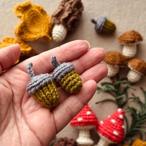 Pode incluir: Bolotas de croch&ecirc; feitas &agrave; m&atilde;o em amarelo e cinza, seguradas em uma m&atilde;o. Outros itens de croch&ecirc; incluem cogumelos, folhas e uma trufa. Os cogumelos t&ecirc;m chap&eacute;us marrons e caules brancos. As folhas s&atilde;o amarelo mostarda.