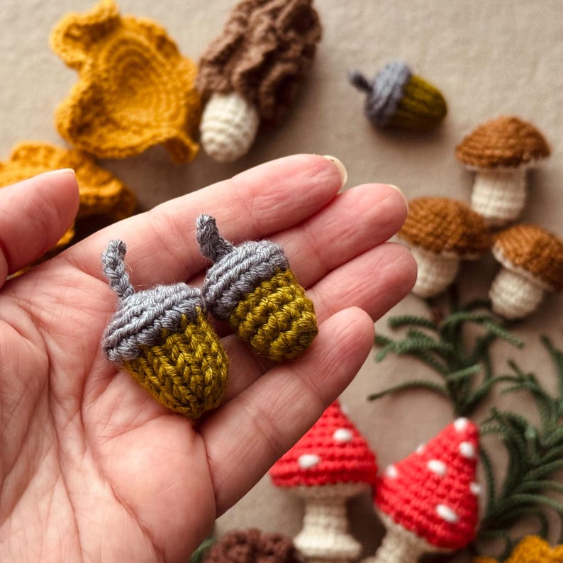 Pode incluir: Bolotas de croch&ecirc; feitas &agrave; m&atilde;o em amarelo e cinza, seguradas em uma m&atilde;o. Outros itens de croch&ecirc; incluem cogumelos, folhas e uma trufa. Os cogumelos t&ecirc;m chap&eacute;us marrons e caules brancos. As folhas s&atilde;o amarelo mostarda.