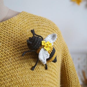 May include: A crocheted bee brooch featuring a black head, black and yellow striped body, white wings, and small yellow flower accents. The bee is attached to a mustard yellow knitted sweater, showcasing detailed yarn work and textured stitching.