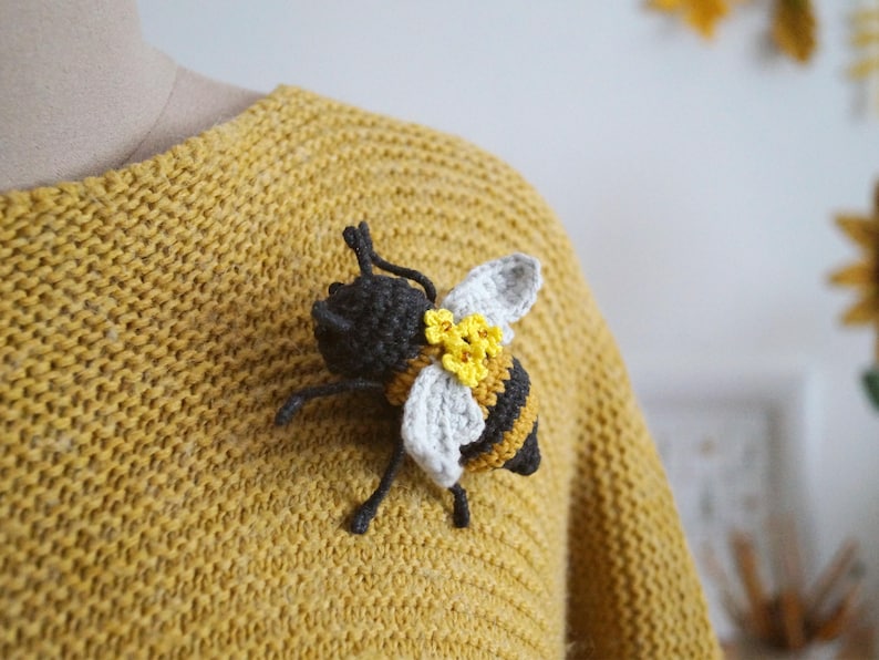 May include: A crocheted bee brooch featuring a black head, black and yellow striped body, white wings, and small yellow flower accents. The bee is attached to a mustard yellow knitted sweater, showcasing detailed yarn work and textured stitching.