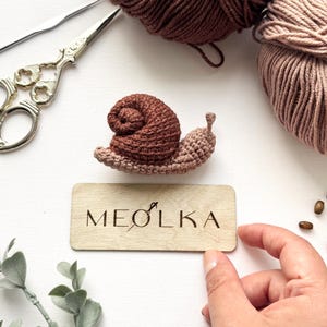 May include: A small crocheted snail with a dark brown shell and a light brown body, positioned above a rectangular wooden plaque engraved with the text MEOLKA, accompanied by two balls of yarn, metal scissors, and small wooden beads.
