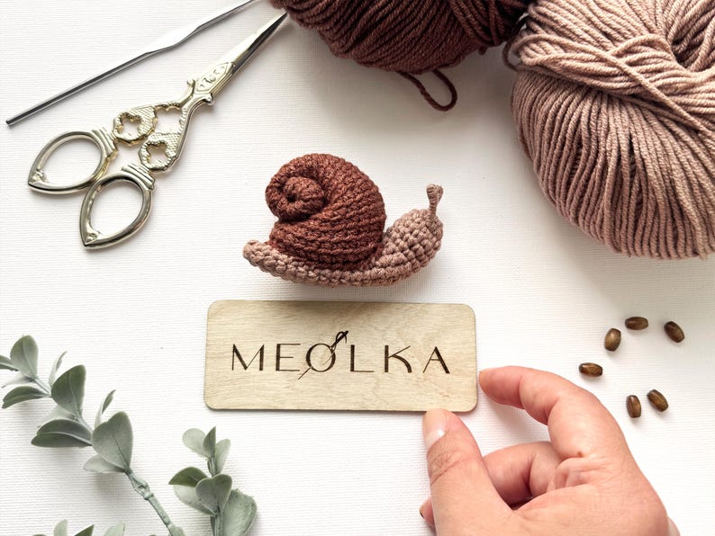 May include: A small crocheted snail with a dark brown shell and a light brown body, positioned above a rectangular wooden plaque engraved with the text MEOLKA, accompanied by two balls of yarn, metal scissors, and small wooden beads.