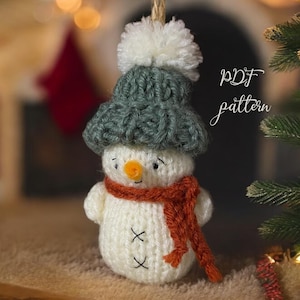 May include: A handmade snowman ornament with a white body, a green knit hat with a white pom-pom, and an orange scarf. The snowman has a carrot nose and black stitched eyes and mouth. The text "PDF pattern" is written in cursive.