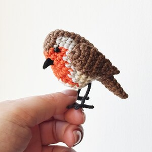 May include: A handcrafted crochet bird figurine. The bird features a brown and white head, a vibrant orange chest, and a black beak. The bird is perched on black wire legs, creating a charming, whimsical design.