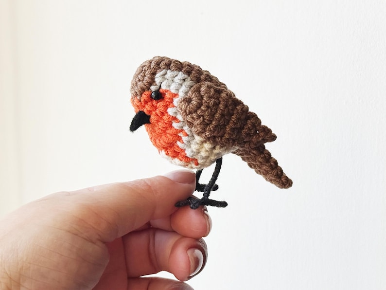 May include: A handcrafted crochet bird figurine. The bird features a brown and white head, a vibrant orange chest, and a black beak. The bird is perched on black wire legs, creating a charming, whimsical design.
